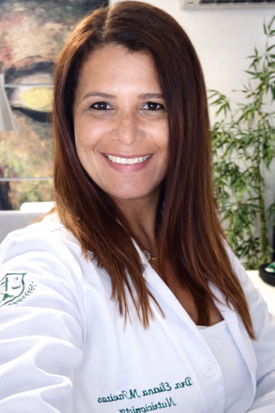 Dra. Eliana Maria de Freitas em seu consultório, de jaleco, sorrindo em frente a uma planta e uma obra na parede.